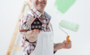 Residential Repainting Painter. Man In White Clothes Painter Holding A Model House And Smiling.